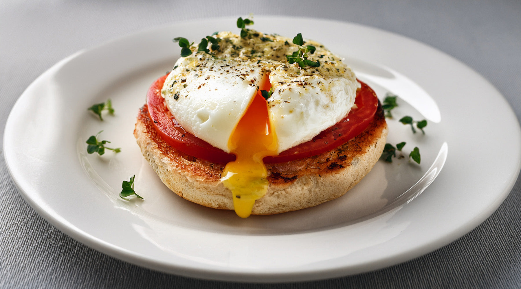 Toasted english muffin, topped with an overeasy egg and a slice of tomato, with herb mayonnaise on top on a white plate